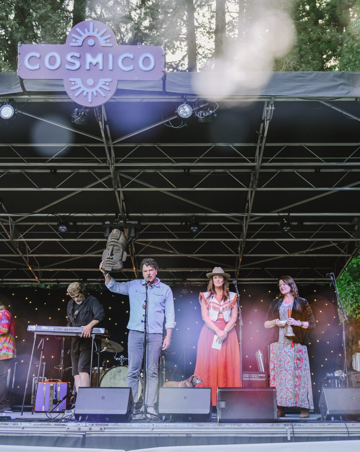 Chris Schultz onstage beneath the Cosmico sign in Sonoma County, holding a carved figure aloft beside the team